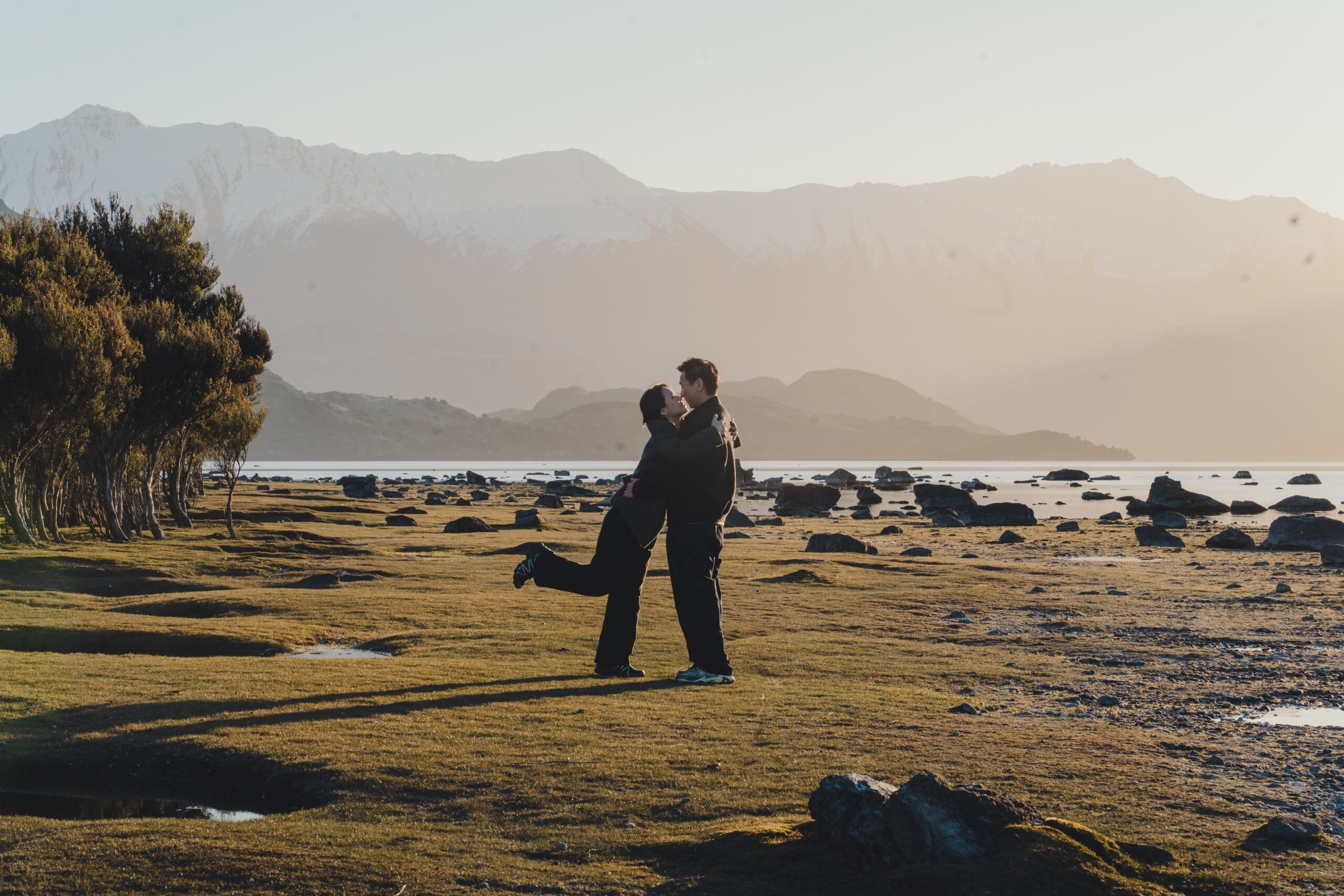 Marcia and Jacky kissing beside Lake Wānaka with Marcia lifting one leg in a playful romantic pose