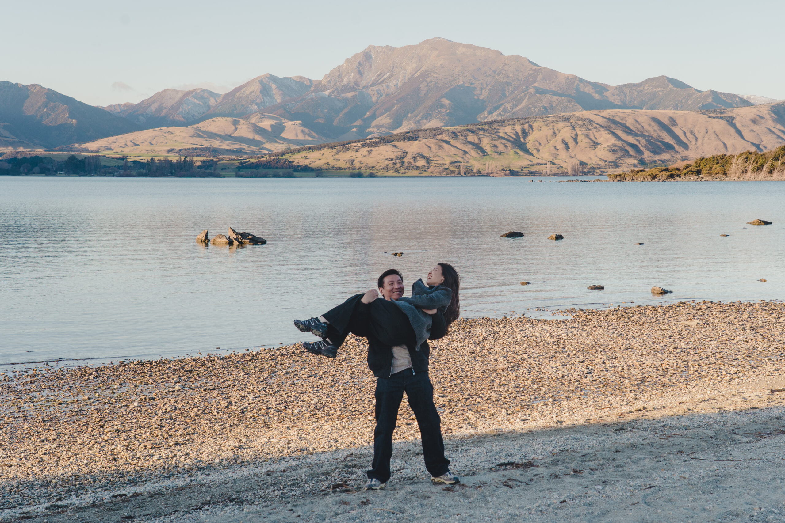 Man lifting fiance up in his arms at the edge of Lake Wanaka. Both laughing
